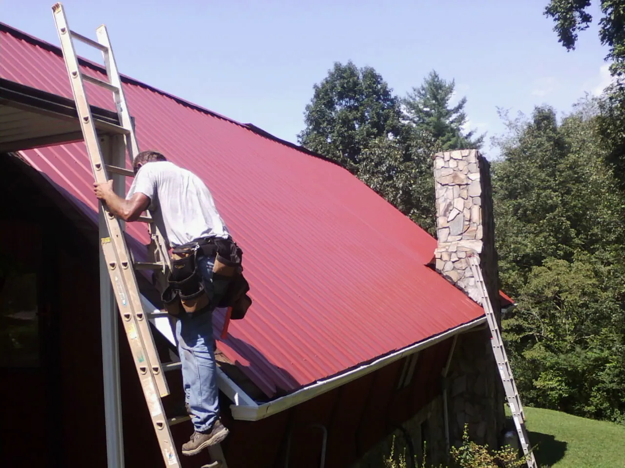 Roofer on a ladder beside a red metal roof and stone chimney — Soffit & Fascia Repair by Roofing Brothers in White Post